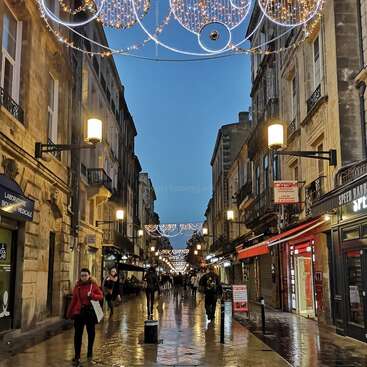 Das Bild zeigt eine nasse Straße, auf der Menschen unterwegs sind, gesäumt von Gebäuden und festlichen Lichtern, vor einem blauen Himmel. Die Atmosphäre ist lebendig und festlich.