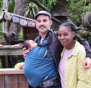 A man carries a sleeping baby in a blue sling while smiling alongside a woman in a yellow jacket. They stand outdoors surrounded by greenery.