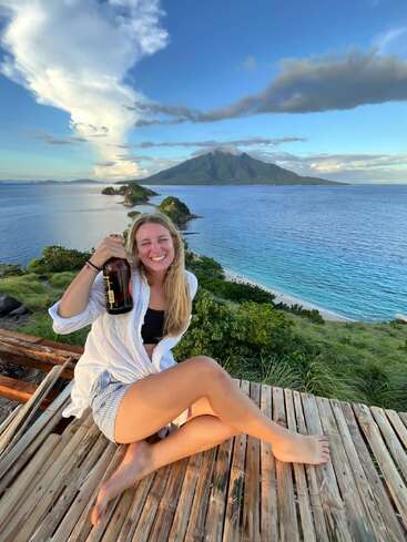 Una mujer sonriente descansa en una terraza de madera, con una botella en la mano, rodeada de una exuberante vegetación, aguas turquesas y una majestuosa isla volcánica al fondo, bajo un cielo azul.