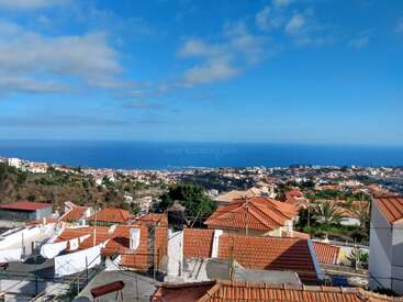 The image shows a coastal town with red-tiled rooftops, green trees, blue sky, scattered clouds, and a stunning view of the vast ocean horizon.