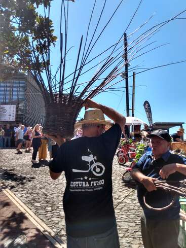 A man wearing a straw hat carries a large basket with long sticks. People gather outside in sunlight, possibly at a market or festival, on cobblestone streets.