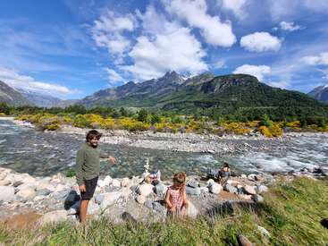 A group of people enjoy a riverside scene, surrounded by rocks, colorful flowers, and lush mountains under a bright, blue sky with fluffy clouds.