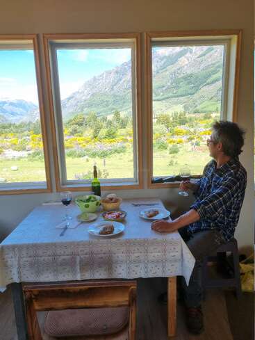 A man sits at a table with food and wine, gazing out large windows at a stunning mountain landscape filled with greenery and wildflowers under blue skies.