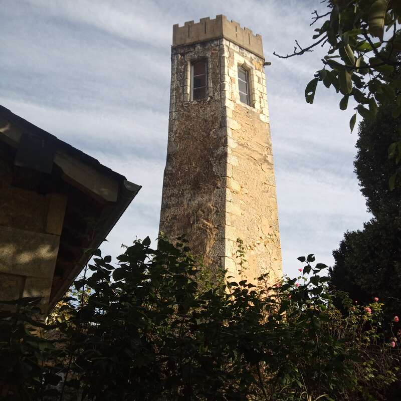 The image depicts a tall stone tower with a window, set against a cloudy sky, surrounded by lush greenery and trees, with a building partially visible on the left.