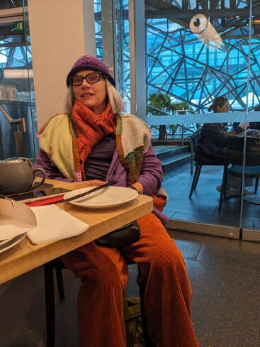Eine Frau in farbenfroher Kleidung, mit Brille und Strickmütze sitzt an einem Cafétisch mit leeren Tellern und Tee, hinter ihr ein moderner Glashintergrund.