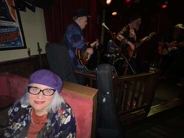 Eine Frau mit weißen Haaren, einer Brille und einer lila Baskenmütze sitzt lächelnd in einem schwach beleuchteten Lokal. Hinter ihr spielt eine Live-Band auf der Bühne.