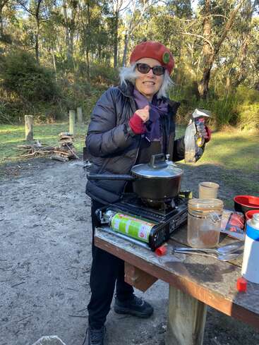 Eine Frau mit einer roten Baskenmütze lächelt, während sie im Freien kocht. Sie steht an einem Tisch mit einem Gasherd, umgeben von Bäumen und Campingausrüstung.