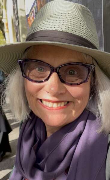 Une femme souriante aux cheveux argentés, portant un chapeau de soleil vert clair, des lunettes noires et une écharpe violette, profite d'une journée ensoleillée à l'extérieur, rayonnant de chaleur et de bonheur.