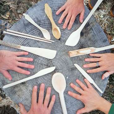 The image depicts a group of people gathered around a tree stump, showcasing their handiwork with various wooden spoons and tools, surrounded by a serene natural setting.