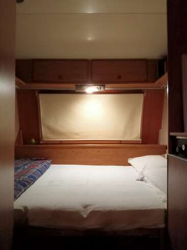 Este quarto aconchegante tem uma cama bem arrumada com lençóis brancos, painéis de madeira, armários suspensos, uma janela coberta por uma cortina bege e uma iluminação suave.