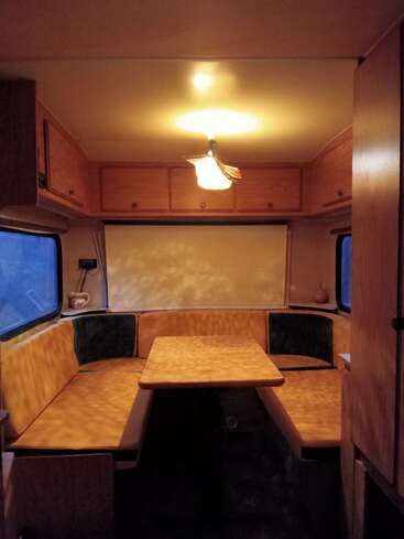 Esta imagem mostra o interior aconchegante de uma van de camping, com uma mesa de madeira, bancos almofadados ao redor, janelas, armários de armazenamento no teto e uma luz de teto que dá um toque aconchegante.