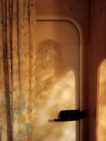 Um quarto mal iluminado tem uma cortina com estampa floral, uma porta fechada com uma trava e um rosto misterioso e fantasmagórico, vagamente visível através do vidro fosco.