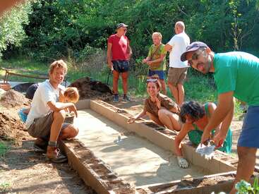 Um grupo de pessoas está trabalhando junto ao ar livre, construindo algo em um ambiente natural. Algumas estão de joelhos, alisando uma superfície arenosa, sorrindo e colaborando.