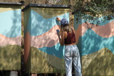 Uma mulher usando um boné e uma blusa regata marrom-avermelhada pinta um mural colorido em estruturas de madeira ao ar livre, segurando o celular com uma mão, cercada pela natureza.