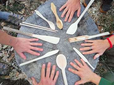 The image depicts a group of people gathered around a tree stump, showcasing their handiwork with various wooden spoons and tools, surrounded by a serene natural setting.