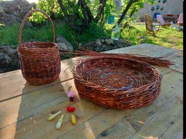 A imagem mostra uma mesa de madeira rústica com duas cestas trançadas, cercada por um ambiente externo sereno com árvores, grama e uma cadeira, evocando uma sensação de tranquilidade e artesanato.