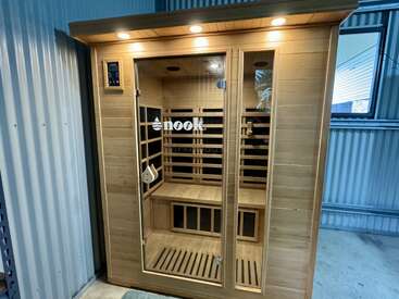 Cette image montre une cabine de sauna infrarouge moderne en bois, appelée "nook", avec une porte en verre, un banc intérieur, des lumières LED et un panneau de commande numérique à l'intérieur d'une pièce aux parois métalliques.