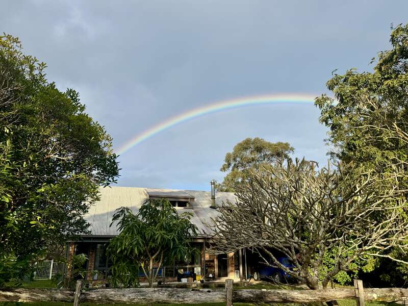 Une maison rustique au toit métallique est entourée d'arbres verdoyants. Au-dessus, un arc-en-ciel vibrant s'étend dans le ciel nuageux, créant une scène paisible.