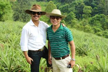 Two men are standing in a green field wearing hats and sunglasses. Both are smiling, with one arm around each other, enjoying the natural outdoor scenery.