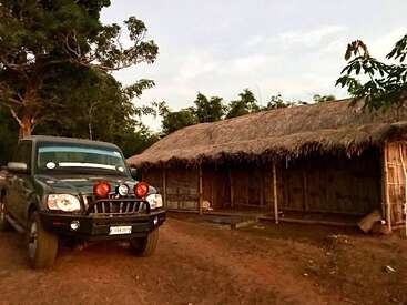 A rugged black SUV is parked on a dirt ground near a long thatched-roof hut, surrounded by trees, suggesting a rural or forest setting at dusk.