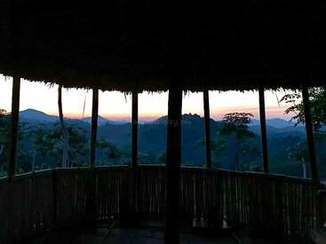 This image shows a bamboo hut at sunset. Silhouetted trees and mountains are visible in the background, framed by the open wooden structure's roof and railings.