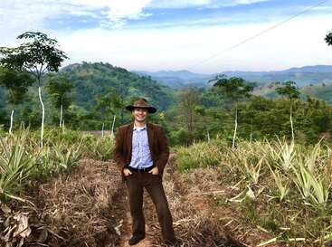 A man in a brown suit and hat stands on a farm path surrounded by lush green hills, young trees, and pineapple plants under a blue sky.
