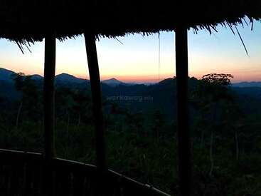 A tranquil view from a rustic wooden hut at dusk, overlooking misty mountains and silhouetted trees beneath a beautifully colored, peaceful sunset sky. Nature at rest.