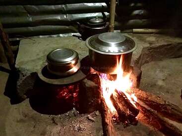 A traditional kitchen setup with metal pots cooking over an open wood fire, surrounded by stone and metal supports, creating a rustic, warm, and homely atmosphere.