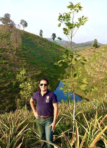 A person stands smiling on a hillside with sunglasses, surrounded by plants and a young tree, overlooking a small river winding through green, scenic hills.