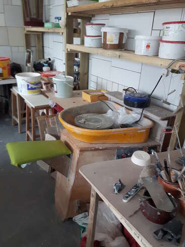 Um estúdio de cerâmica com um torno amarelo, assento verde, prateleiras com baldes de materiais, várias ferramentas, pincéis e suprimentos espalhados por mesas e prateleiras.