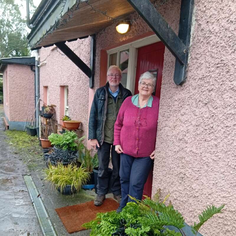 Um casal de idosos está na entrada de uma casa rosa, cercada por vasos de plantas e vegetação, sorrindo calorosamente sob a luz da varanda em um dia chuvoso.