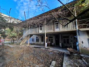 This image shows an old, two-story house with a weathered exterior, surrounded by leafless trees. A person stands by the entrance, and scattered leaves cover the ground.