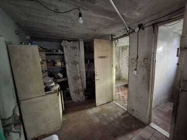 This image shows a dimly lit, cluttered basement or storage room with old cabinets, shelves, exposed wiring, a bare lightbulb, and stained walls and floors.