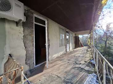 This image shows a weathered balcony with peeling floor, rusted railing, and cracked walls. An open door, windows, and tree branches complete the abandoned atmosphere.