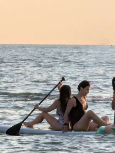 Two women sit back-to-back on a paddleboard in the sea, enjoying a calm sunset. One holds a paddle as gentle waves surround them.