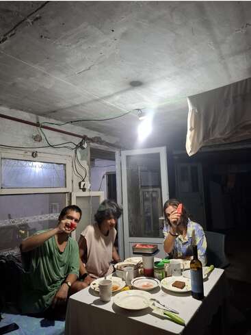 Three people sit around a dinner table in a rustic, dimly-lit room. Plates and cups scatter the table. Two hold up watermelon slices, smiling. Cozy, casual atmosphere.
