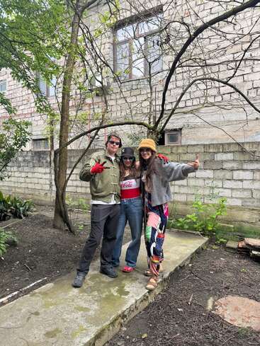 Drei Freunde posieren fröhlich draußen auf einem Betonweg, in bunten Outfits und mit Hüten. Sie stehen neben einem Baum vor einem Backsteingebäude und zeigen das Peace-Zeichen.