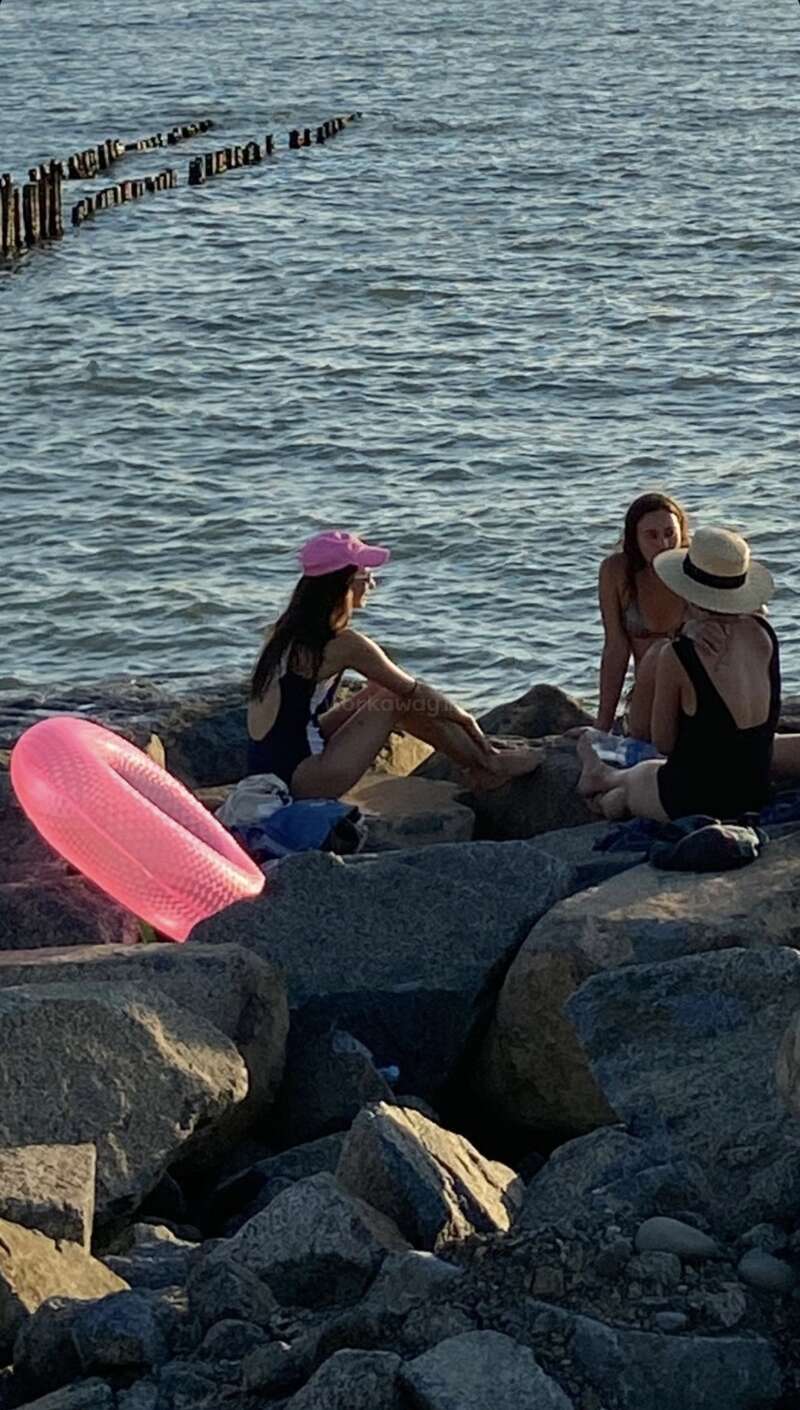 Three women in swimsuits sit on rocks by the water, talking. A bright pink inflatable floats nearby, with worn wooden posts in the background. Peaceful scene.