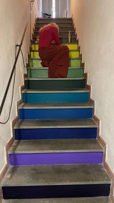 Jemand mit langen Haaren sitzt auf einer regenbogenfarbenen Treppe und malt oder arbeitet. Die Stufen haben verschiedene leuchtende Farben. Daneben steht eine Wasserflasche. Drinnen.