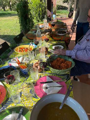 Ein bunter Tisch im Freien ist gedeckt für ein Essen mit bunten Tellern, Weingläsern, Salaten, Brot und Leuten, die ein sonniges, entspanntes Beisammensein in der Natur genießen.