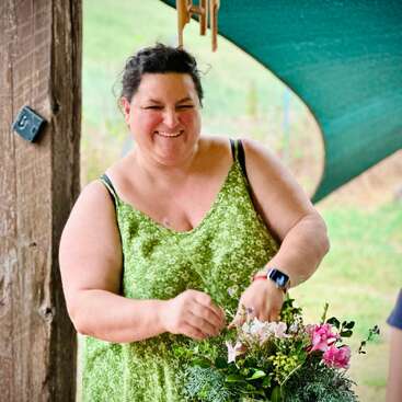 A smiling woman in a green dress arranges a colorful bouquet of flowers outdoors, standing beside a wooden post under a shaded area, looking cheerful and content.