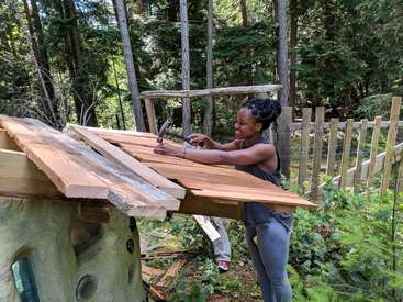 Das Bild zeigt eine Frau, die in einem Waldgebiet an einer Holzkonstruktion arbeitet und mit einem Hammer Bretter an einem Rahmen befestigt.