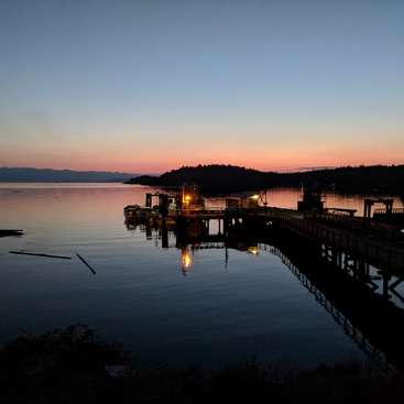 La imagen representa una serena escena en un muelle al atardecer, con una tranquila masa de agua, un muelle con una barca y un pequeño edificio, sobre un fondo montañoso.