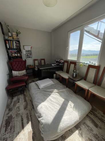 Ein gemütlicher, sonniger Raum mit einem Einzelbett auf dem Boden, umgeben von Stühlen, einem Schreibtisch, einem Bücherregal, einem roten Sessel und großen Fenstern mit Blick auf die malerische Natur.