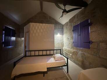 Cette image montre une chambre à coucher rustique, aux murs de pierre, avec un seul cadre de lit en métal, des fenêtres à volets violets, un ventilateur au plafond, un éclairage minimal et des meubles simples.