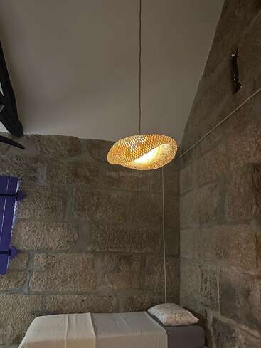Une chambre confortable avec des murs en pierre, un lit simple avec une literie blanche et un oreiller à motifs, et une lampe tressée unique suspendue au plafond, illuminée.