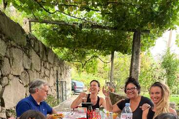 Un groupe de personnes est assis à une table en plein air sous un auvent de vigne verte, dégustant de la nourriture, des boissons et des rires à côté d'un mur de pierre rustique.