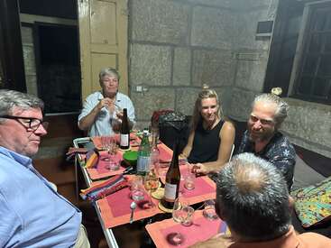 Cinq personnes sont assises autour d'une table dans un intérieur confortable. Elles semblent prendre un verre et discuter, avec des bouteilles, des verres et de la nourriture sur la table.
