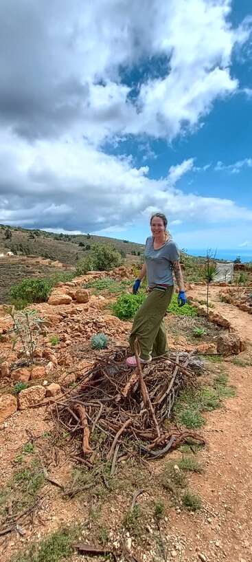 Una mujer sonriente, con guantes y ropa informal, está de pie sobre un montón de ramas en un paisaje rural y rocoso, bajo un cielo azul parcialmente nublado.