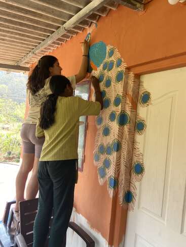 Deux femmes peignent une fresque représentant un paon sur un mur orange à l'extérieur. L'une d'elles est debout sur une chaise, toutes deux concentrées sur l'ajout de couleurs vives et de détails.